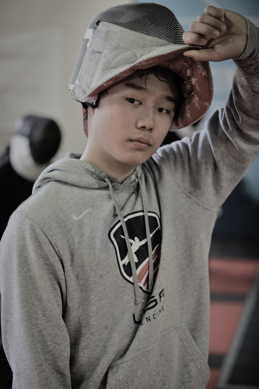 A boy in a saber fencing mask wearing a USA fencing sweatshirt in a saber private lesson