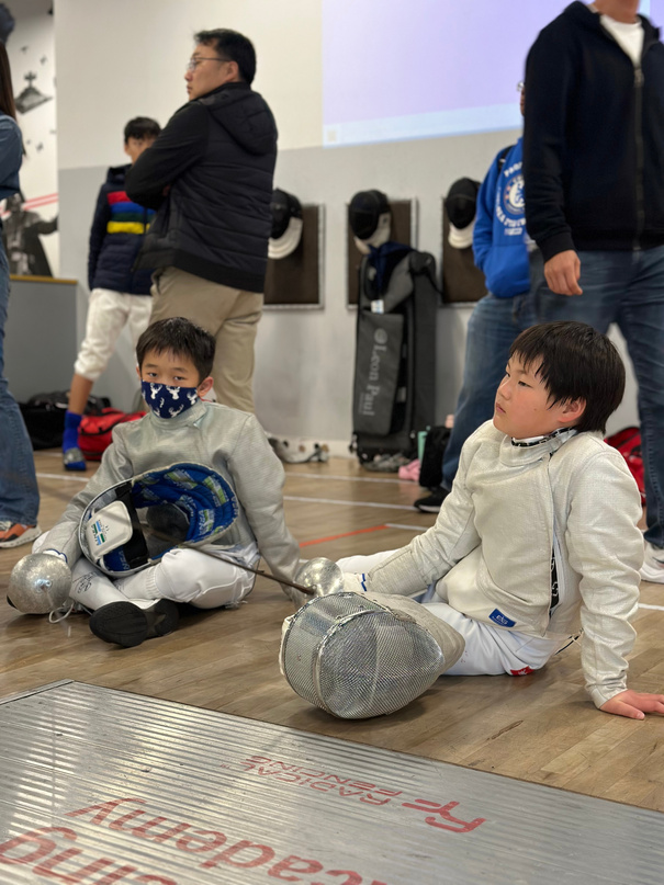 Two young boys in fencing gear sitting on the floor during a regional fencing tournament waiting for their tournament. 