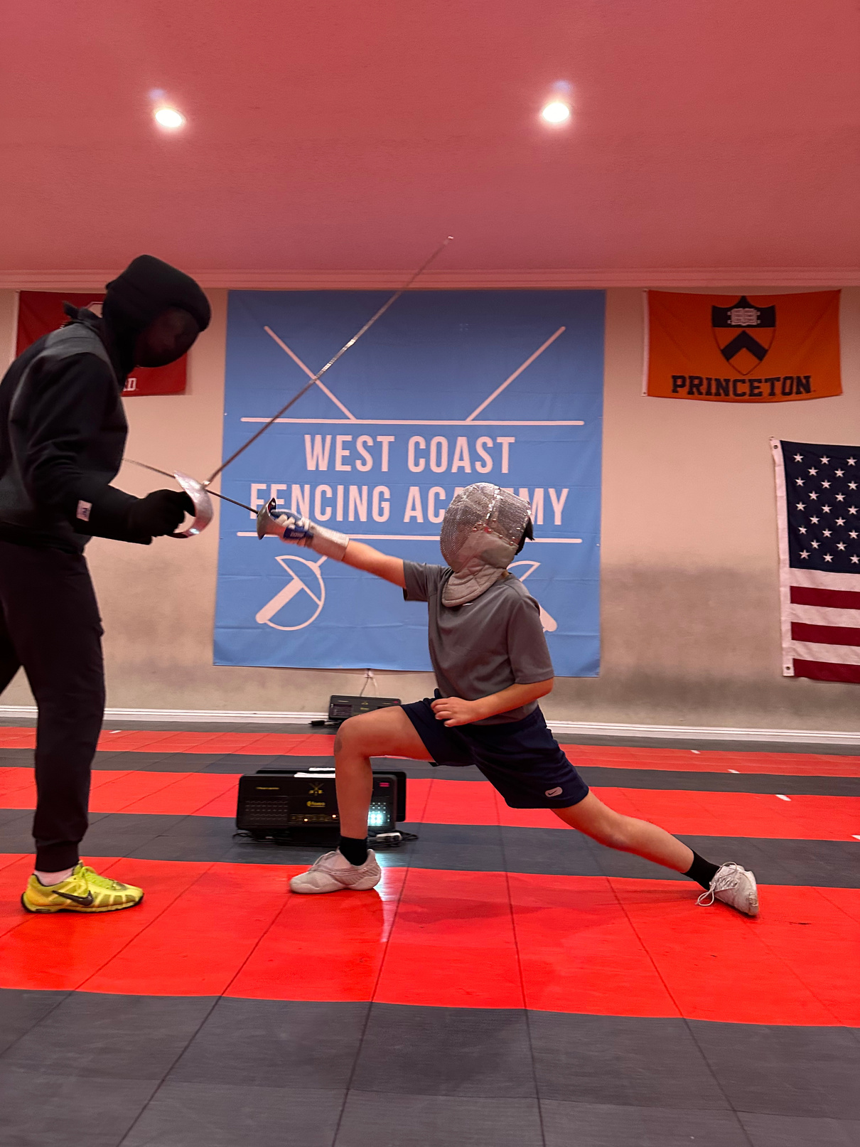 Coach Artem having a private lesson with a young boy who is wearing fencing mask and holding a sword in a fencing lunge in front of the west coast fencing academy flag. 