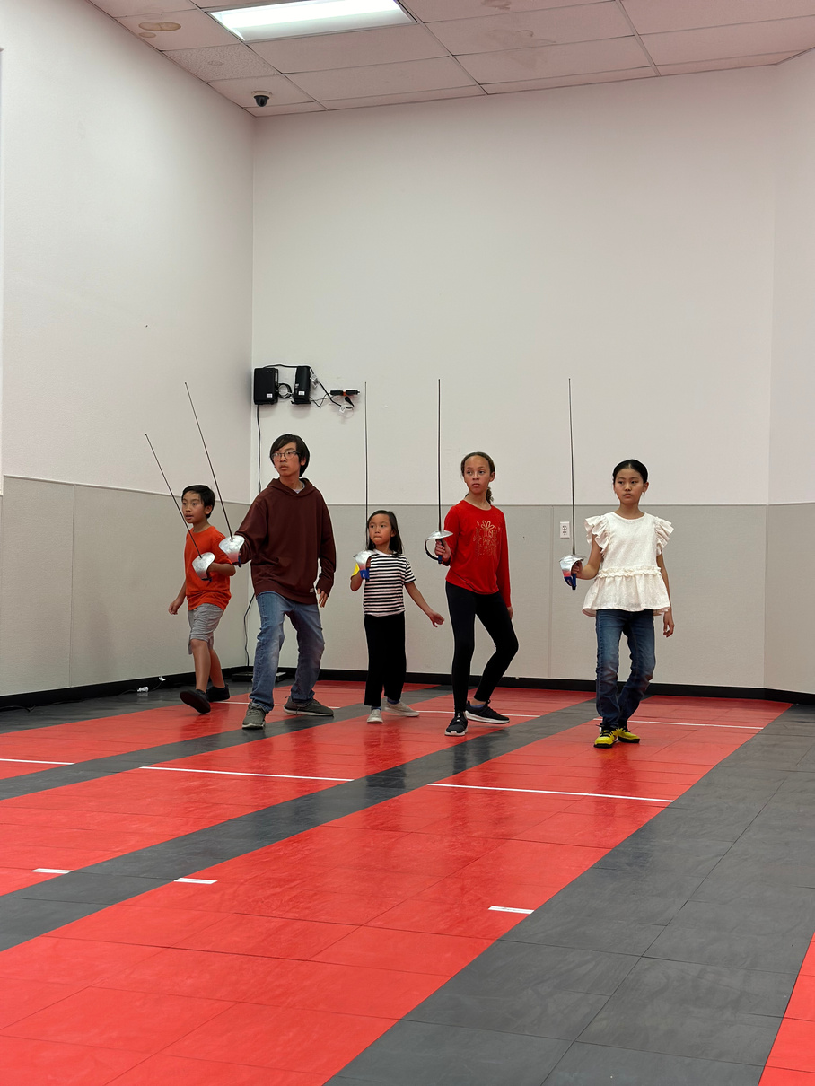 A group of 8 - 12 year old beginner saber fencers learning footwork holding a saber. 
