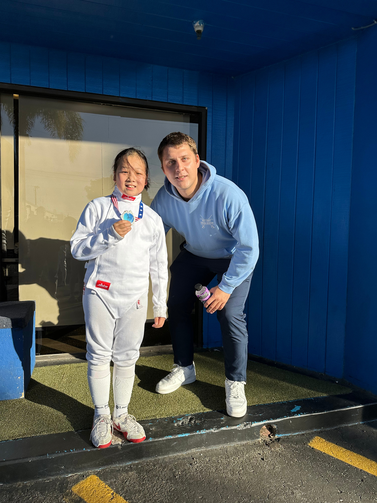 coach mike kishchenkov posing with girl saber fencing student holding a medal she won at her fencing tournament
