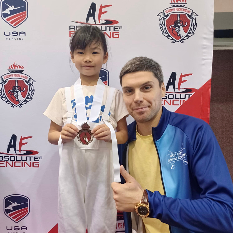 coach artem zanin with a thumb up pose in west coast fencing academy jacket next to a young girl in fencing gear wearing a medal she won from her fencing tournament